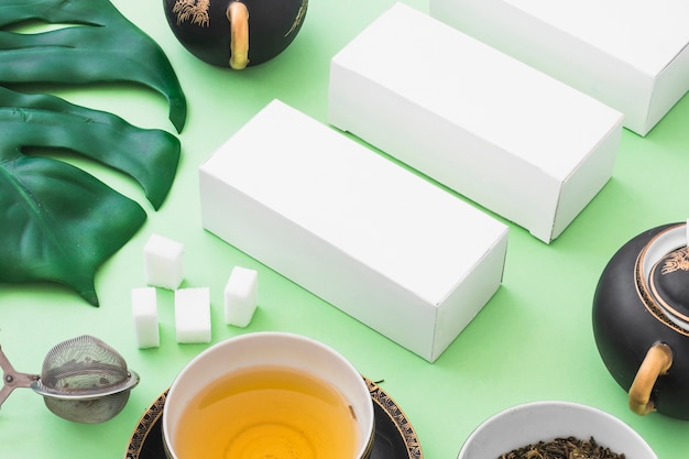 Té de hierbas, cubos de azúcar, colador de té y cajas blancas sobre fondo verde