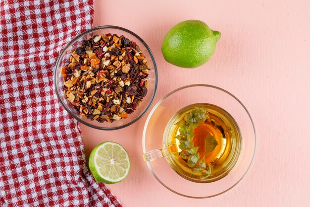 Té de hierbas en una taza de vidrio con limas, hierbas secas planas sobre una toalla rosa y de cocina