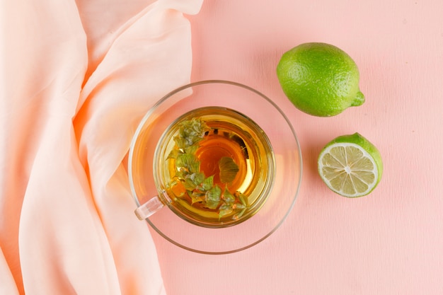 Té de hierbas en una taza de vidrio con limas planas sobre rosa y textil