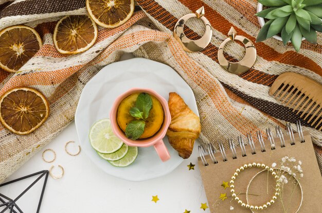 Té de limón a base de hierbas con croissant y accesorios femeninos sobre mantel sobre fondo blanco