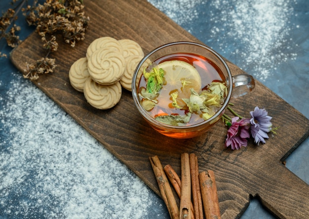 Té de limón con galletas, hierbas secas, palitos de canela en azul sucio y tabla de cortar, plano.