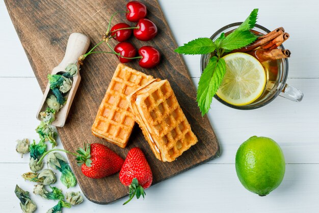 Té con limón, gofres, hierbas secas, frutas, menta, canela en una taza de madera y tabla de cortar, plano.