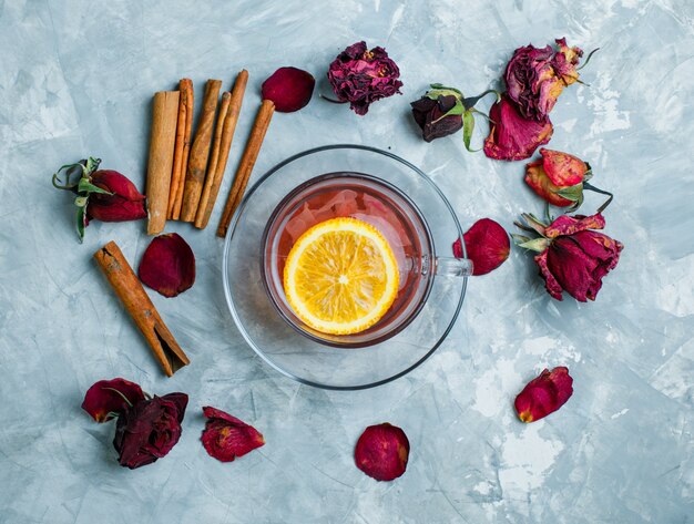 Té de limón con palitos de canela, rosas secas en una taza sobre fondo azul sucio, vista superior.