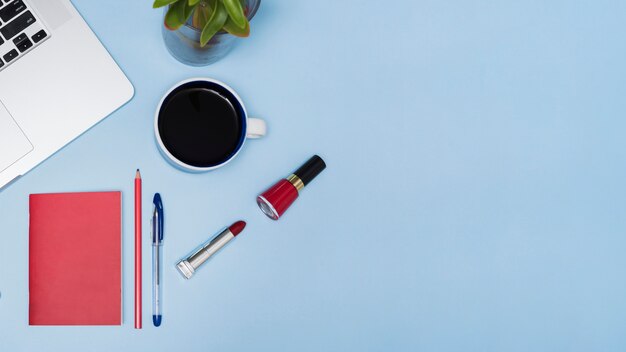 Te negro ordenador portátil; planta; papelería y lápices labiales sobre fondo azul