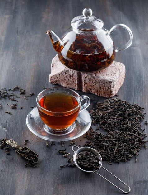 Té negro en tetera y taza con té seco, vista de ángulo alto de ladrillo sobre una superficie de madera