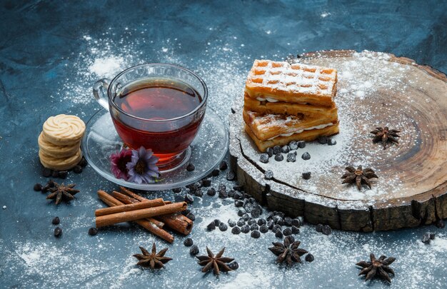 Té en una taza con gofres, galletas, choco chips, especias vista de ángulo alto en grunge azul y tablero de madera