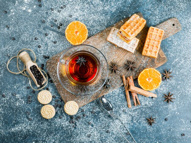 Té en una taza con gofres, galletas, especias, choco chips, colador, vista superior naranja en azul y tabla de cortar