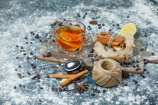 Té en una taza con harina, chips de chocolate, colador, especias, limón, vista de ángulo alto sobre hormigón y tabla de cortar