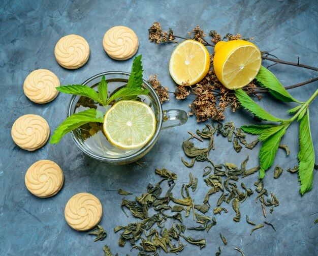 Té en una taza con limón, galletas, menta, hierbas secas planas sobre una superficie azul