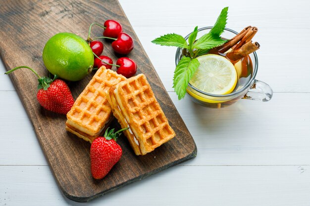 Té en una taza con limón, gofres, fresas, cerezas, menta, canela, plano sobre madera y tabla de cortar