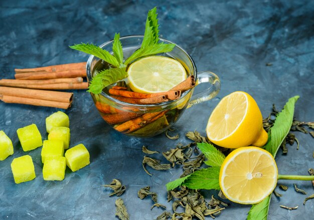 Té en una taza con limón, menta, palitos de canela, terrones de azúcar vista de ángulo alto sobre una superficie azul