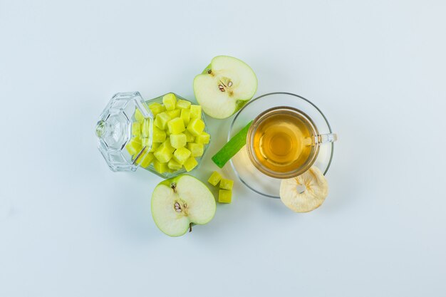 Té en una taza con manzana, frutos secos, terrones de azúcar, caramelos sobre un fondo blanco.