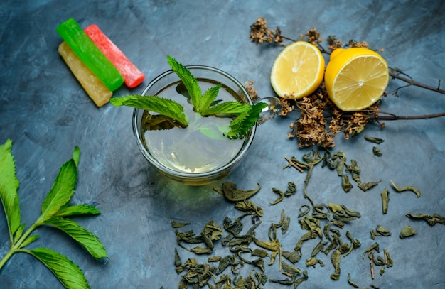 Té en una taza con menta, hierbas secas, limón, caramelos planos sobre una superficie azul oscuro