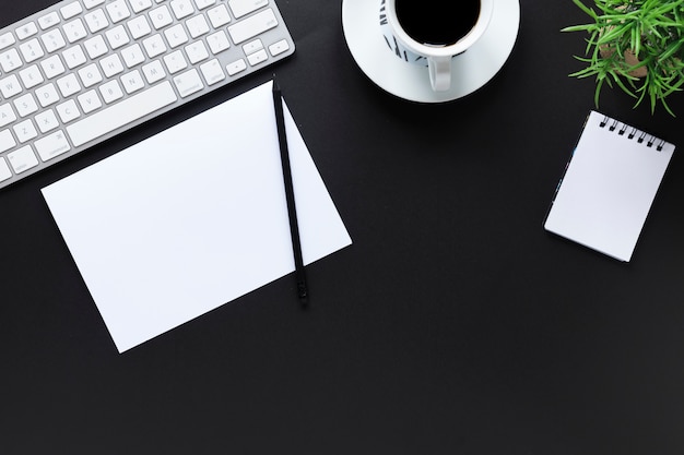 Teclado blanco; papel; lápiz; taza de café; Bloc de notas espiral y planta de maceta en fondo negro