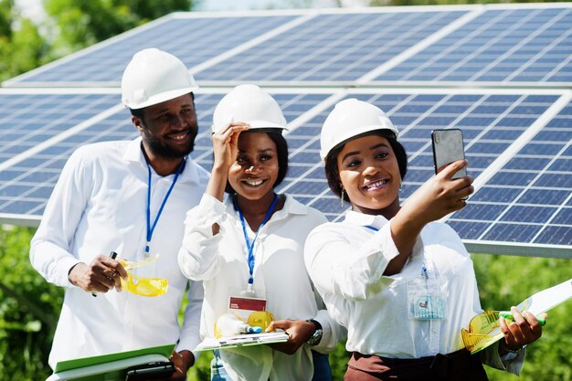 Técnico afroamericano verifica el mantenimiento de los paneles solares Grupo de tres ingenieros negros reunidos en la estación solar Hacer selfie por teléfono