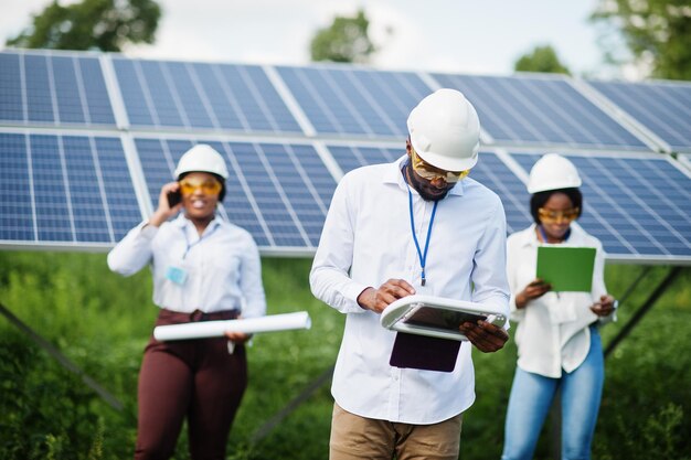 Técnico afroamericano verifica el mantenimiento de los paneles solares Grupo de tres ingenieros negros reunidos en la estación solar