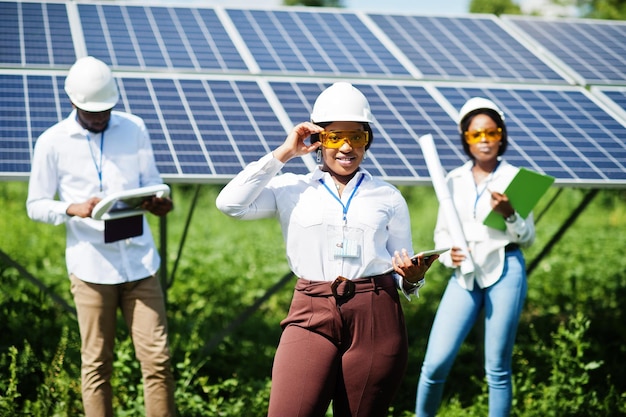 Técnico afroamericano verifica el mantenimiento de los paneles solares Grupo de tres ingenieros negros reunidos en la estación solar