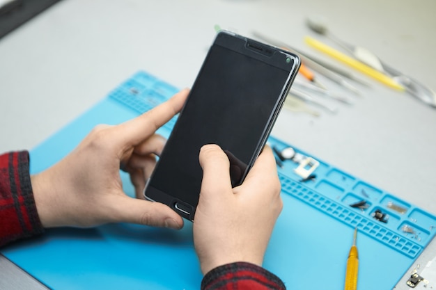 Foto gratuita técnico con camisa a cuadros sentado en su lugar de trabajo, encendiendo el teléfono inteligente en sus manos para encontrar fallas