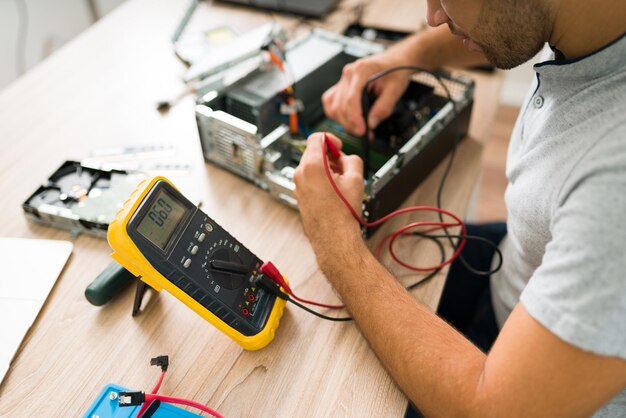 Técnico que usa un multímetro para medir el voltaje de una computadora rota. Joven revisando las conexiones de la computadora en el taller de reparación