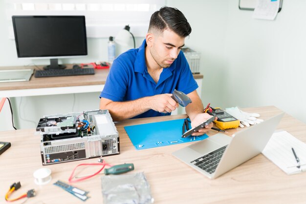 Técnico trabajador escaneando el código de barras de un viejo componente roto para buscar una pieza de repuesto en el taller de reparación