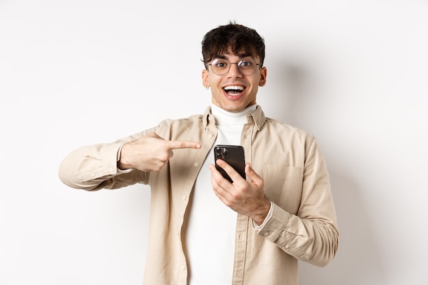 Tecnología y concepto de compras online. Chico hipster feliz apuntando con el dedo a la pantalla del teléfono inteligente y sonriendo, mirando la promoción de la aplicación, de pie sobre fondo blanco