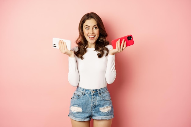 Foto gratuita tecnología, concepto de promoción de primavera. mujer glamorosa con estilo sosteniendo dos teléfonos inteligentes, teléfonos móviles en ambas manos, sonriendo complacida, comprando un nuevo teléfono celular, fondo rosa