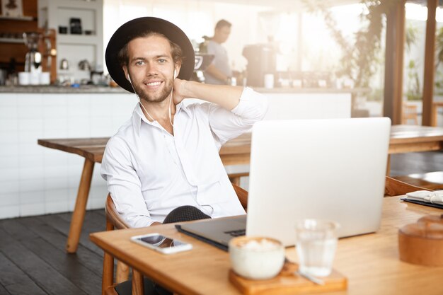 La tecnología hace la vida más fácil. Hombre barbudo de moda en auriculares usando conexión inalámbrica gratuita a internet en su computadora portátil, escuchando música o audiolibros en línea durante el almuerzo en el interior de la cafetería moderna