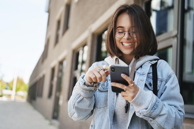 Tecnología juvenil y concepto de primavera Una mujer joven y bonita alegre con gafas, una chaqueta de mezclilla, riendo y sonriendo, mirando la pantalla del teléfono móvil, envía mensajes de texto divertidos mientras está de pie en la calle