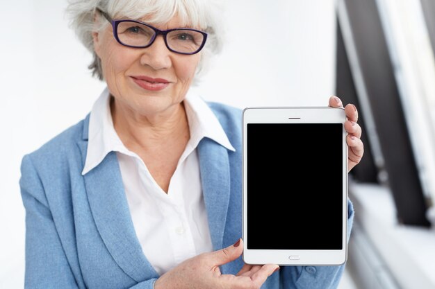 Tecnología moderna, envejecimiento y concepto de comunicación en línea. Atractiva empresaria madura feliz de sesenta años en stlyish anteojos sonriendo y sosteniendo tableta digital con pantalla en blanco