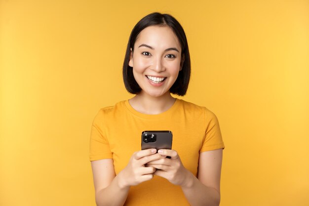 Tecnología Mujer asiática sonriente usando un teléfono móvil sosteniendo un teléfono inteligente en las manos de pie en una camiseta contra un fondo amarillo