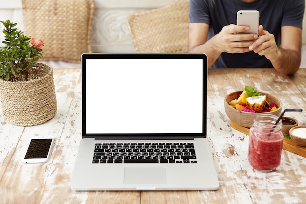 Tecnología, negocios, comunicación, personas y publicidad. Vista frontal del lugar de trabajo de diseño de madera con portátil abierto con pantalla en blanco, teléfono celular, vaso de batido y tazón de frutas.