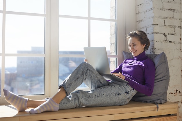 Tecnología, ocio, diversión y relax. Encantadora joven vistiendo elegantes jeans y cuello alto relajándose en el alféizar de la ventana en casa con una computadora portátil en su regazo