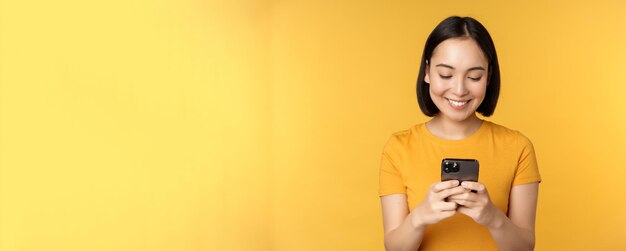 Tecnología sonriente mujer asiática usando teléfono móvil con teléfono inteligente en las manos de pie en camiseta aga...