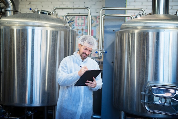 Tecnólogo sonriente tomando notas en la línea de producción de la fábrica satisfecho con los resultados