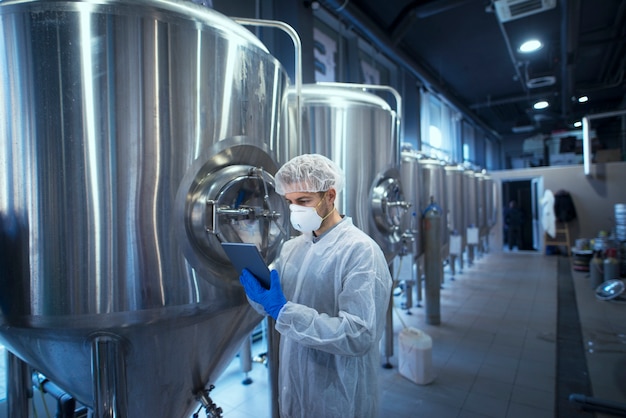 Foto gratuita tecnólogo trabajador de fábrica en uniforme protector con redecilla y máscara que controla la producción de alimentos en la tableta