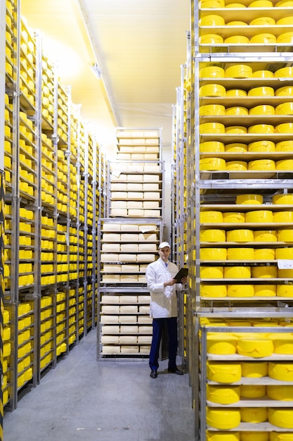 Tecnólogo en una túnica blanca que sostiene una tableta para registros y se encuentra cerca de los estantes con quesos Producción de productos de queso Hombre en la tienda de quesos