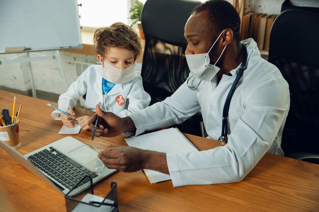 Teenboy caucásico como médico consultando, dando recomendaciones, tratando. Pequeño doctor durante la discusión, estudiando con un colega mayor. Concepto de infancia, emociones humanas, salud, medicina.