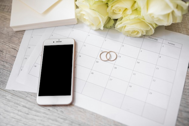 Teléfono inteligente; anillos de boda; Sobre y rosas en el calendario sobre el tablón de madera.