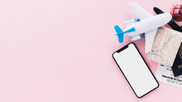Teléfono inteligente con pantalla en blanco con pasaporte de viaje; mapa; Entradas; avión de juguete y gafas de sol sobre fondo rosa