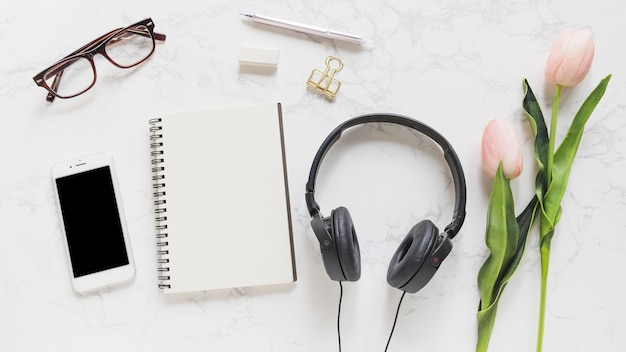 Teléfono móvil; los anteojos; cuaderno; papelería; auriculares y tulipanes de color rosa sobre fondo de mármol