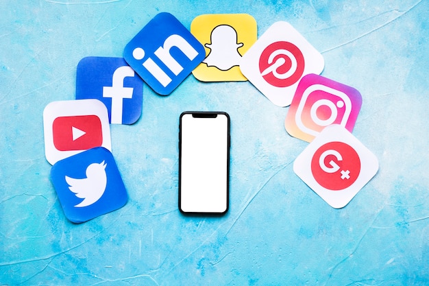 Teléfono móvil en blanco con varios iconos de papel de redes sociales sobre fondo azul