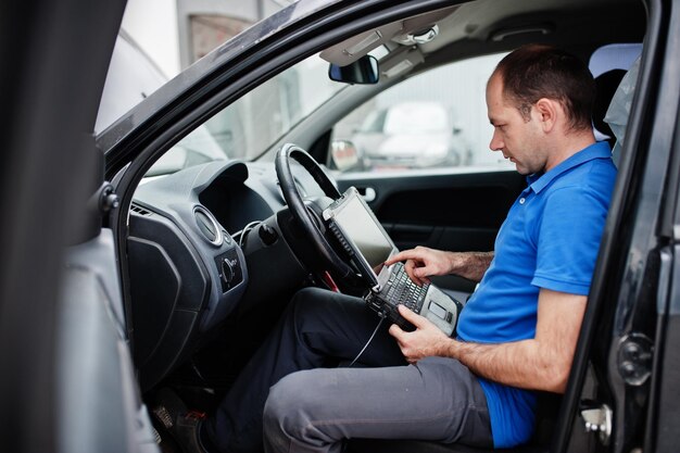 Tema de reparación y mantenimiento de automóviles Mecánico eléctrico en uniforme que trabaja en el servicio de automóviles haciendo diagnósticos de automóviles usando un dispositivo obd con una computadora portátil