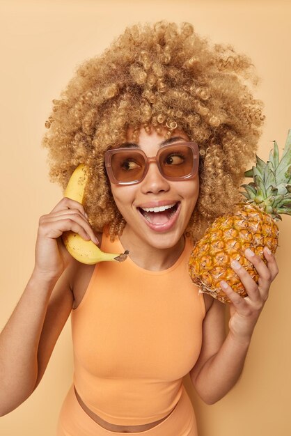 Temporada de frutas tropicales Una mujer alegre con cabello rubio y rizado hace tonterías y finge hablar por teléfono sostiene un plátano cerca de la oreja en una mano piña fresca en la otra usa ropa deportiva y gafas de sol