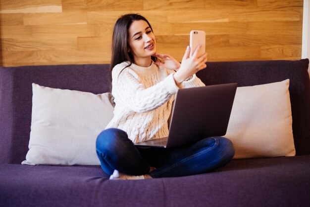 Tender lady está trabajando en una computadora portátil y hablando por teléfono sentada en la cama de la casa