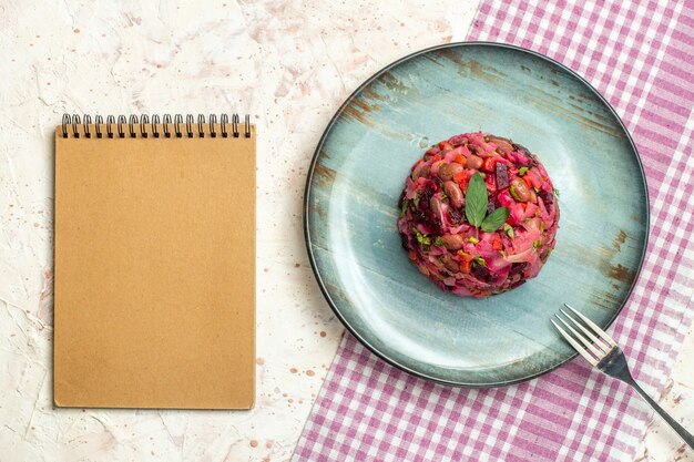 Tenedor de ensalada de vinagreta de vista superior en placa ovalada en cuaderno de mantel a cuadros blanco y morado en mesa gris claro