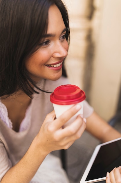 Foto gratuita la tenencia sonriente de la muchacha se lleva la taza de café y la tableta digital a disposición