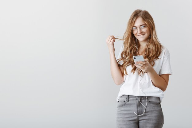 Tener ideas desagradables en mente. Retrato de hermosa estudiante rubia encantadora con gafas, sosteniendo un teléfono inteligente, mordiéndose el labio y jugando con el cabello, sonriendo con curiosidad, coqueteando o en un estado de ánimo romántico