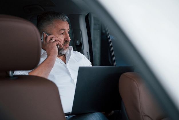 Tener una llamada de negocios sentado en la parte trasera del automóvil con una computadora portátil de color plateado