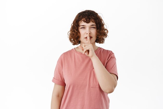 Tener un secreto. Linda chica rizada callando y guiñando un ojo, escondiendo algo, callando tímidamente, dile que se calle, de pie en una camiseta en blanco