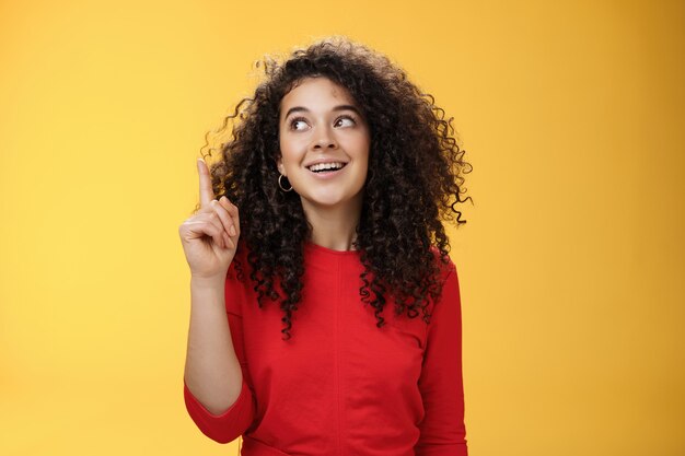 Tengo idea emocionado mujer atractiva feliz con el pelo rizado en vestido rojo levantando el dedo índice en eurek ...